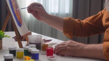 An elderly male artist paints a colorful artwork at home. His hands skillfully work on a canvas with various paints and brushes.