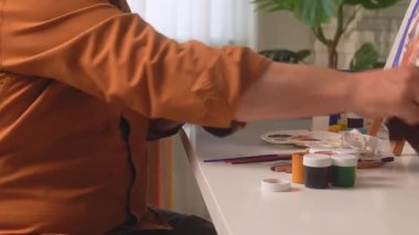 Close up of a man in a brown shirt wiping his hands with a cloth after finishing painting. Art supplies, brushes, and paint jars on a desk. Concept of creativity, art, and relaxation.