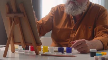 An elderly male artist is painting at home, surrounded by art supplies. His work focuses on intricate details.