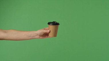 Bir kadın eli çerçeveye uzanır, kahverengi bir kahve fincanını yeşil arka planda siyah bir kapakla tutar..