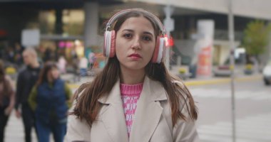 Pembe kulaklık takan ve bej ceketli genç bir kadın kalabalık bir caddede insanlarla yürüyor..