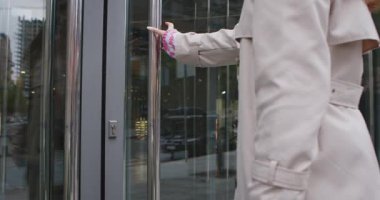 Trençkot giyen bir kadın cam bir kapı açar ve şehir ortamında modern bir binaya girer. Yavaş çekim.