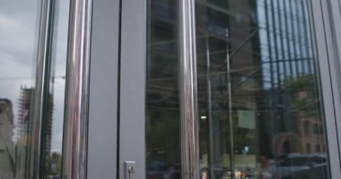 Yağmurluk giymiş bir kadın cam bir kapı açar ve şehir ortamında modern bir binadan çıkar. Büyük bir şehirde bir alışveriş merkezi. Yavaş çekim.