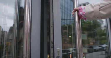 Trençkot giyen bir kadın cam bir kapı açar ve şehir ortamında modern bir binaya girer. Yavaş çekim.