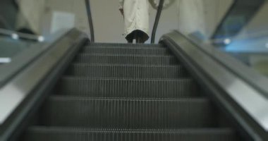 Uzun bej ceketli ve siyah çizmeli biri modern bir alışveriş merkezinde yürüyen merdivene çıkıyor. Yavaş çekim.