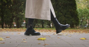 Siyah çizmeleri, bej paltosu ve yere düşmüş yaprakları olan bir adamın parka girerken yakın çekimi..