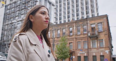 Bej ceketli genç bir kadın, şehirdeki eski ve modern binaların önünde duruyor..