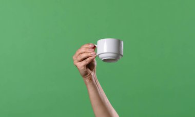 Yeşil krom arka planda beyaz kahve fincanı tutan bir kadın eli. Ofis, iş, eğitim, kafe veya kahve endüstrisiyle ilgili tasarımlar için çekilen.