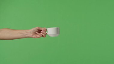 Yeşil krom arka planda beyaz kahve fincanı tutan bir kadın eli. Ofis, iş, eğitim, kafe veya kahve endüstrisiyle ilgili tasarımlar için çekilen.