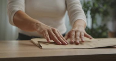 Bir kadın, kağıt bir torbayı ahşap bir masada katlıyor. Basit dekorlu parlak bir oda ve arka planda yeşil bir bitki..