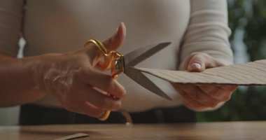 Bir masa üzerinde kahverengi kıvrımlı karton kesmek için altın makas kullanan kadın ellerini gösteren yakın plan bir resim.