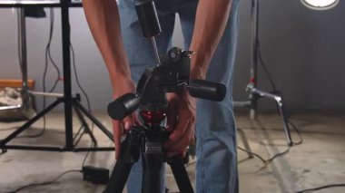 Bir video stüdyosundaki tripodun kafasını ışıklandırma ve ekipman zeminine karşı sıkılaştıran ve ayarlayan bir adama yaklaş. Çekim sırasında kamerayı kurmaya hazırlanılıyor.