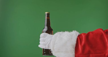 Noel Baba, beyaz kürk manşetli ve eldivenli kırmızı bir elbise içinde yeşil arka planda izole edilmiş koyu renkli bir bira şişesi tutuyor. Noel afişleri, kartlar ve tatil reklamları için mükemmel..