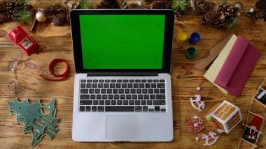 Yeşil renkli ekranı olan bir dizüstü bilgisayarın üst görüntüsü Noel süslemeleri olan ahşap bir masanın üzerinde duruyor. Yeni yıl kutlamalarına hazırlanılıyor..