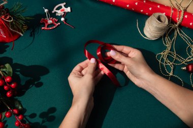 Bayram hediyesi paketi için kırmızı kurdele hazırlayan kadın elleri, Noel süslemeleri, kozalaklar, böğürtlenler ve parlak güneş ışığı altında rulo kağıtlarla çevrili..
