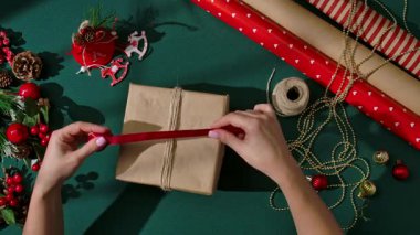 Kadın elinin hediyeyi kahverengi kağıda sardığı ve kırmızı saten kurdeleyi yeşil arka plana bağladığı en iyi manzara. Noel süslemeleriyle çevrili rahat ve şenlikli bir tatil ortamı yaratıyor..