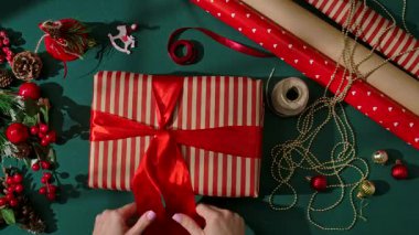 Kırmızı saten bir kurdeleyi çizgili bir hediye kutusuna bağlayan bir kadın eli, makas, sicim ve Noel sanatları için yeşil bir arka planda süslemeler..