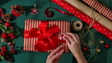Güzel paketlenmiş bir Noel hediyesini parlak kırmızı saten bir fiyonkla Kraft kağıdına sarmış kadın ellerinin üst görüntüsü. Kutlama dekorasyonları hediyeyi sarıp sıcak ve neşeli bir tatil ortamı yaratıyor..