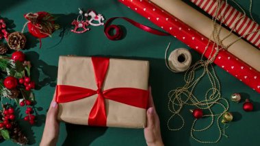 Kırmızı saten kurdele yay ayarlayan kadın eller Kraft kağıdına sarılı Noel hediyesi, şenlikli süslemeler ve yeşil bir arka planda malzeme sarma.