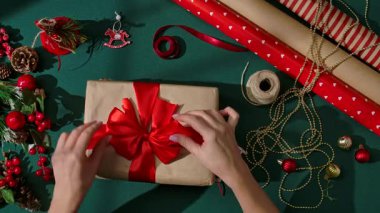 Kırmızı saten kurdele yay ayarlayan kadın eller Kraft kağıdına sarılı Noel hediyesi, şenlikli süslemeler ve yeşil bir arka planda malzeme sarma.