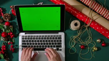 Koyu yeşil şenlik arka planında krom yeşil ekranlı dizüstü bilgisayarda yazı yazan elleri ambalaj malzemeleri ve dekorasyonlarla çevrili. Tatil alışverişi ya da tasarım için mükemmel bir düzen..