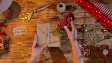 Kraft kağıdına sarılı, sicimle bağlanmış, köknar dalları ve kırmızı böğürtlenlerle süslenmiş, kırsal ahşap bir masa üzerinde makas ve kurdele kullanan bir kadın eli. Konforlu el yapımı tatil paketi.