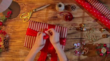 Kırmızı saten bir kurdeleyi çizgili bir hediye kutusuna bağlayan bir kadın eli, makas, sicim ve kırsal bir ahşap masa üzerindeki süslemeler Noel zanaatı için..