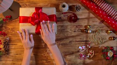 Noel hediyelerini kırmızı kurdelelerle istifleyen kadın elleri ve ahşap bir masada Mutlu Noeller kartı. Dekorasyonlu, el yapımı şenlikli ambalaj.