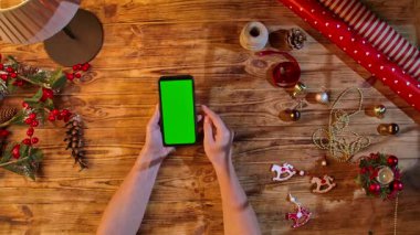 Noel süslemeleri ve ambalaj malzemeleriyle çevrili kırsal ahşap bir masa üzerinde yeşil ekranlı akıllı telefonu araklayan eller, şenlikli alışveriş veya tasarım düzeni için ideal..