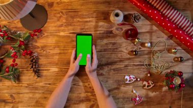 Noel süslemeleri ve ambalaj malzemeleriyle çevrili kırsal ahşap bir masa üzerinde yeşil ekranlı bir akıllı telefonu daktilo eden eller, festival alışverişi veya tasarım düzeni için ideal.