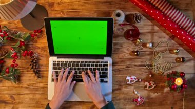Ellerin dizüstü bilgisayarda yazışının üst görüntüsü. Kromatik yeşil bir ekranla ahşap bir masada. Etrafı ambalaj malzemeleri ve dekorasyonlarla çevrili. Tatil alışverişi ya da tasarım için mükemmel bir düzen..