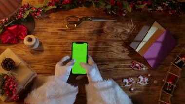 Noel Baba 'nın eldivenli eldiveni, ahşap bir masada yeşil krom bir ekranla akıllı bir telefona yazı yazarken üst görüntüsü. Yılbaşı ve Noel kutlamalarına hazırlanılıyor.