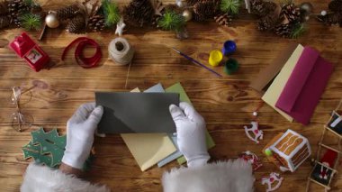 Noel Baba 'nın eldiven taktığı el yazısı bir Noel mektubunu taşralı ahşap bir masada kurdeleler, boyalar ve süslemeler ile el ele tutuşarak, yazı yazmak için yeri olan samimi bir tatil sahnesi oluşturduğu görünümü..