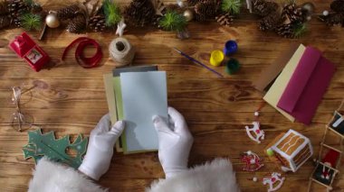 Noel Baba 'nın eldivenli ellerinin rengarenk Noel kartlarını kırsal bir masa üzerinde kurdeleler, pullar ve paketlenmiş bir hediyeyle seçmesinin üst görüntüsü. Sanatçılık için şölen sahnesi.