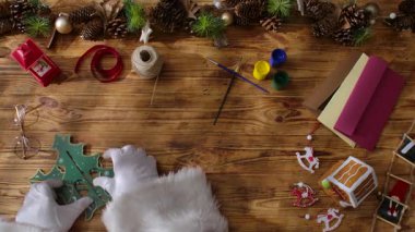 Noel Baba 'nın eldivenli üst görüntüsü boyalar, kurdeleler ve makaslarla süslü süslemeler yaratıyor. Kırsal bir ahşap masanın yanında sarılı bir hediye..