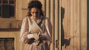 Tarihi giysiler içinde zarif bir kadın antik bir binanın devasa, yıpranmış sütunları arasında duruyor ve sıcak akşam ışığında bir mektup okuyor..