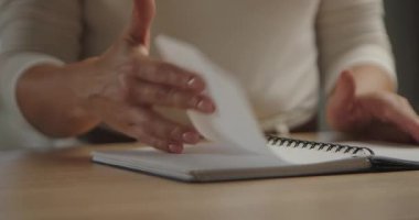 Ahşap bir masanın üzerinde boş bir defterin sayfalarını çeviren bir kadının elleri samimi ve yaratıcı bir iş anını yakalıyor. Yavaş çekim.