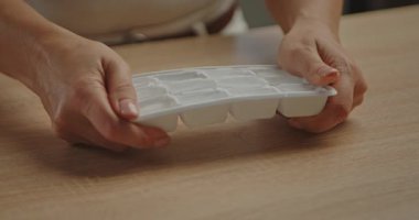 Donmuş küpleri mutfak ışıklarının altındaki ahşap bir masaya koymak için plastik bir buz kalıbını çeviren bir kadının ellerine yakın çekim. Yavaş çekim.