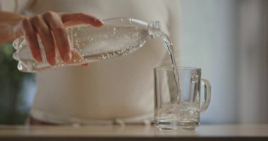 Bir kadın eli, plastik bir şişeden temiz köpüklü suyu ahşap bir masaya yerleştirilmiş şeffaf bir cama döküyor. Yavaş çekim.