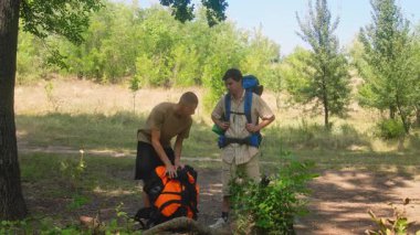 İki genç sırt çantası gezisi sırasında dinleniyor. Biri su içerken diğeri sırt çantasıyla devrilmiş bir ağaca yaslanıyor. Çevrelerinde yeşil bir yaz ormanı var..