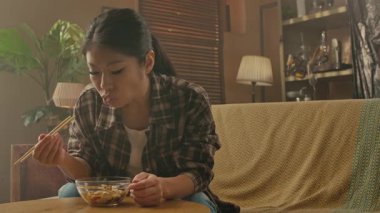 A Korean woman is sitting on a sofa, enjoying a meal of Asian food using chopsticks in a cozy home setting.