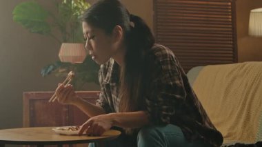 A Korean woman is sitting at a table enjoying traditional Asian cuisine with chopsticks in a cozy home setting.