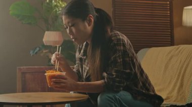 A Korean woman sits comfortably at home, savoring Asian food with chopsticks in a cozy setting.