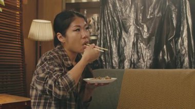 A Korean woman is sitting on a cozy couch, enjoying a plate of delicious Asian food with chopsticks in a warm setting.