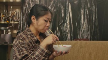 A Korean woman using chopsticks to eat traditional Asian food while sitting in a cozy room.