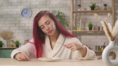 Kızıl saçlı genç bir kadın mutfak masasında beyaz bir bornozla oturuyor, tabakta yemek kesiyor. Ev mutfağında kahvaltı, yemek yeme ritüeli..