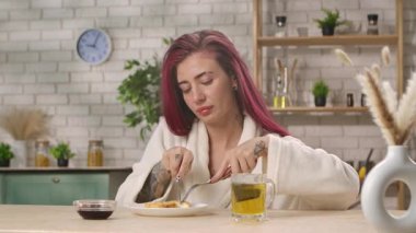 Beyaz bornozlu bir kadın mutfak masasında oturmuş sıcak bir evde bitki çayıyla kahvaltı yapıyor. Ev mutfağında kahvaltı, yemek yeme ritüeli..