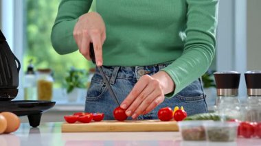 Yeşil kazaklı bir kadın mutfak tezgahında kiraz domatesleri kesiyor, evde kahvaltı hazırlıyor..