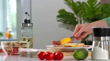 Yeşil kazaklı bir kadın kahvaltı hazırlar ve tabağa kızarmış sosis, pastırma ve mantar koyar. Etrafında çeşitli malzemeler var. Ev mutfağında lezzetli yemekler hazırlıyorum..