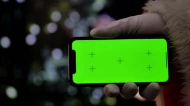 Noel Baba 'nın elinde beyaz bir eldiven, şöminenin önünde yeşil ekran krom anahtarı ve Noel ağacı olan bir akıllı telefon var. Dijital ya da temalı modeller için sıcak bir festival atmosferi. Yavaş çekim.